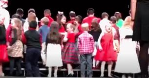 Des enfants s'alignent sur scène alors qu'ils tournent autours d'une jeune fille en robe rouge ce qui fait diviser le public